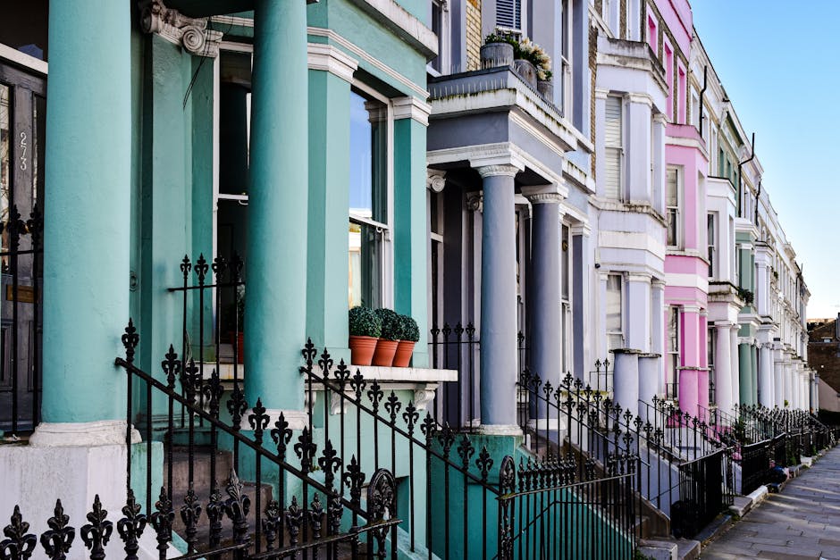 A row of multi-storey terraced houses painted in bright, distinct colours including beige, deep blue, vibrant green, and pink, situated along a street with clear, sunny weather. Each building features