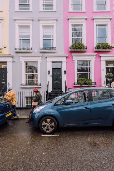 A quiet residential street in Notting Hill with a cobblestone pathway lined by terraced houses, some of which feature white painted facades and brick exteriors. Several potted plants and small trees a