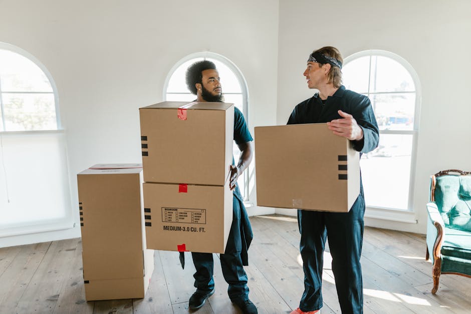 Two professional removalists from Removal Van Notting Hill are preparing for home relocation inside a bright room with arched windows allowing natural light. The worker on the left, with curly hair and a beard, is holding two stacked cardboard boxes, one of which has a label indicating a medium size 3 cubic foot capacity. The other worker, wearing a black headband and dark clothing, is carrying a single cardboard box and appears to be engaged in conversation. Several other boxes, made of cardboard and covered with packing tape, are placed on the wooden floor, some on the left and others in the background. The room features a vintage green armchair with wooden arms on the right side, near the window, and the flooring is made of light-toned wood planks. This scene depicts the logistics of packing and loading items in preparation for a structured move, typical of services offered by Removal Van Notting Hill during house removals or furniture transport in the Notting Hill area.