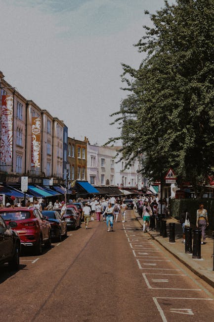 Image shows a busy urban street scene in Notting Hill with a narrow, cobbled roadway lined by parked cars on both sides. Pedestrians are visible walking along the pavement, some carrying shopping bags, while others stand or move across the street. Traditional Victorian-style buildings with colourful facades and awnings house shops and cafes, with some storefronts displaying signage. On the right, a large tree extends overhead, providing partial shade. The scene captures the typical lively atmosphere of a neighbourhood known for its market and vibrant community. This environment reflects the context of logistical planning for house removals or moving services, such as those provided by Removal Van Notting Hill, involving careful loading and transport of household furniture, boxes, and appliances in preparation for a home relocation process.