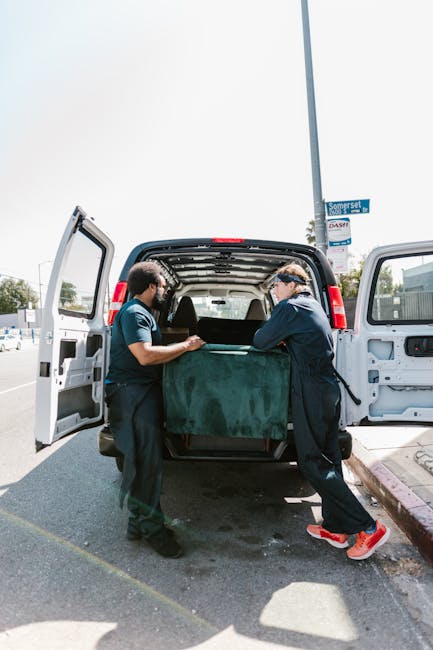 Two professional removalists from Removal Van Notting Hill are preparing for home relocation inside a bright room with arched windows allowing natural light. The worker on the left, with curly hair and a beard, is holding two stacked cardboard boxes, one of which has a label indicating a medium size 3 cubic foot capacity. The other worker, wearing a black headband and dark clothing, is carrying a single cardboard box and appears to be engaged in conversation. Several other boxes, made of cardboard and covered with packing tape, are placed on the wooden floor, some on the left and others in the background. The room features a vintage green armchair with wooden arms on the right side, near the window, and the flooring is made of light-toned wood planks. This scene depicts the logistics of packing and loading items in preparation for a structured move, typical of services offered by Removal Van Notting Hill during house removals or furniture transport in the Notting Hill area.