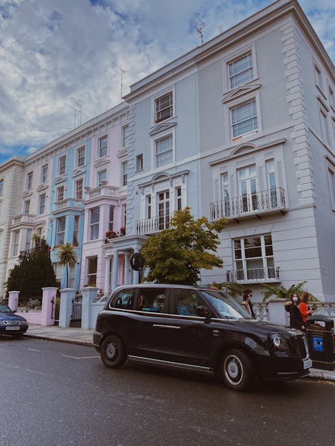 A busy city street scene in Notting Hill during daylight, with pedestrians walking along the pavement and crossing the road. Several cars are parked and moving along the road, including a black taxi.