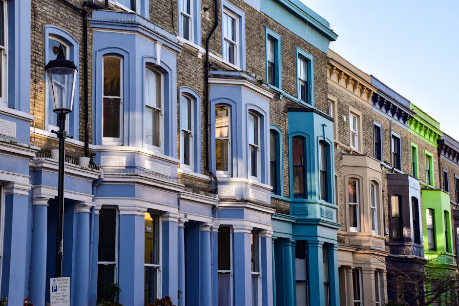 The image depicts a row of colorful, pastel-painted terraced houses on a street in Notting Hill, with some storefronts visible at ground level, including a shop with an awning displaying items outside