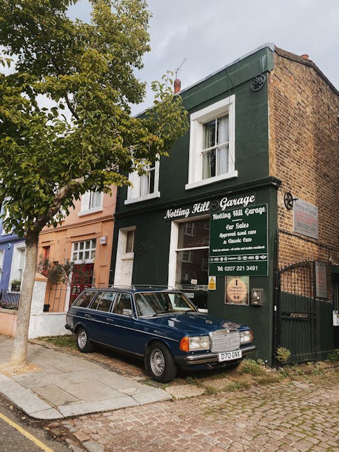 The image shows a two-storey brick building painted dark green, with a sign reading 'Notting Hill Garage' and additional text indicating it offers car sales, repairs, and servicing. In front of the building, a vintage blue station wagon is parked on a cobbled pavement, partially covering the entrance and the shop window. To the left, a tree with green foliage extends its branches over the scene, casting some shade. The surrounding area includes a neighboring pastel pink building with white-framed windows, typical of residential or commercial properties in Notting Hill. The sky is overcast, providing diffuse lighting across the scene. This setting is consistent with a typical residential street where local removals or furniture transport operations could take place, with clear visibility of a vehicle used in home relocation or moving services, supported by Removal Van Notting Hill's general service offerings.